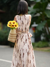 Woman in a sleeveless floral maxi mulberry silk dress holding sunflowers in a woven bag.