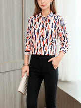 Woman wearing a Women's Geo - Print Casual Silk Blouse with a vibrant pattern and black pants, standing by a window.