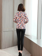 Woman wearing a Women's Geo - Print Casual Silk Blouse with a vibrant pattern and black pants, standing by a window.
