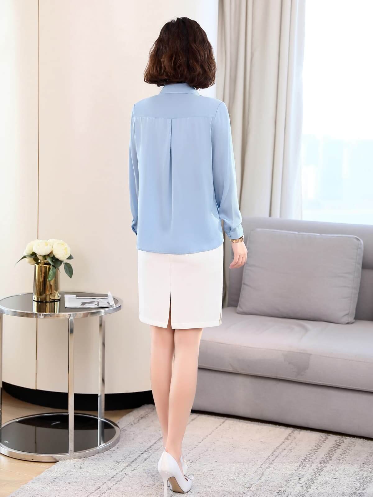 Back view of a woman wearing a light blue versatile mulberry silk blouse and a white skirt, styled in a modern living room.