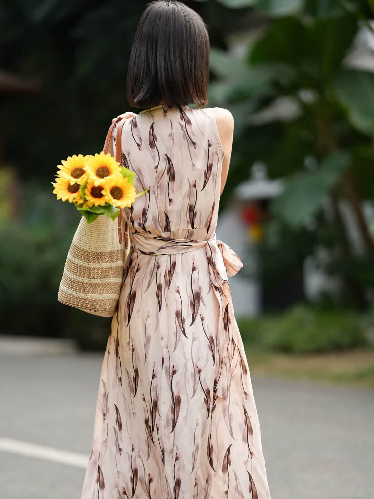 Woman in a sleeveless floral maxi mulberry silk dress holding sunflowers in a woven bag.