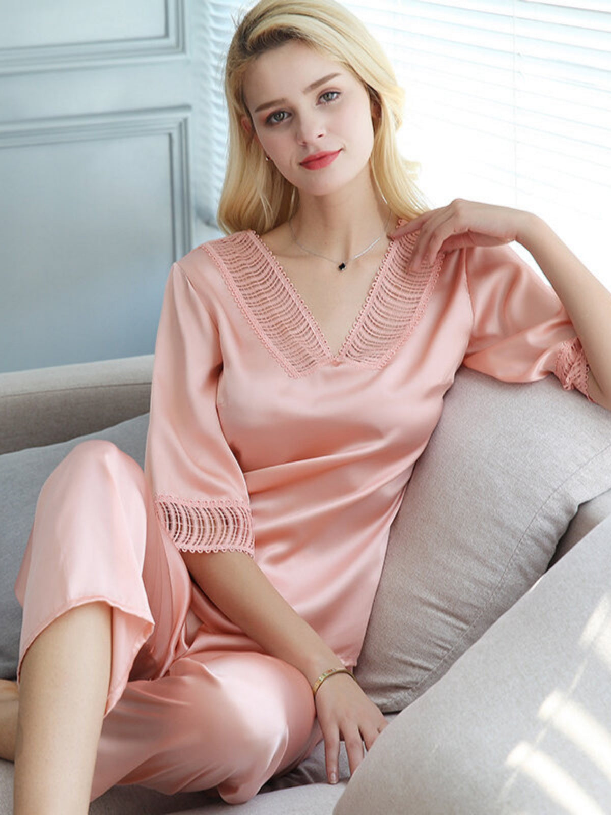 Woman in a pink silk pyjama set sitting on a gray couch in a bright room.